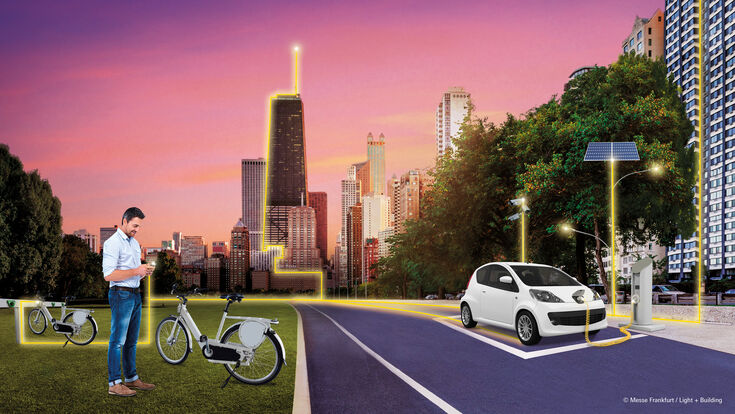 © Messe Frankfurt / Light + Building Mann, der im Gras vor einer Stadt mit Hochhäusern steht, auf der Straße daneben ein Auto, das gerade Strom tankt. Smartphone des Mannes, die Häuser und das Auto sind alle mit einem gelben Strich verbunden.