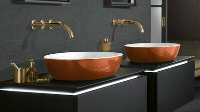 © Villeroy & Boch Zwei Waschschalen, die außen in verbranntem Orange glasiert sind und innen weiß sind über dunkelgrauen Unterschränken vor einer dunkelgrauen Wand und mit messingfarbenen Armaturenn.