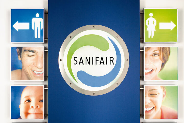 Der Eingang zu einer Sanifair-Toilettenanlage mit einem großen Sanifair-Poster mittig im Bild.