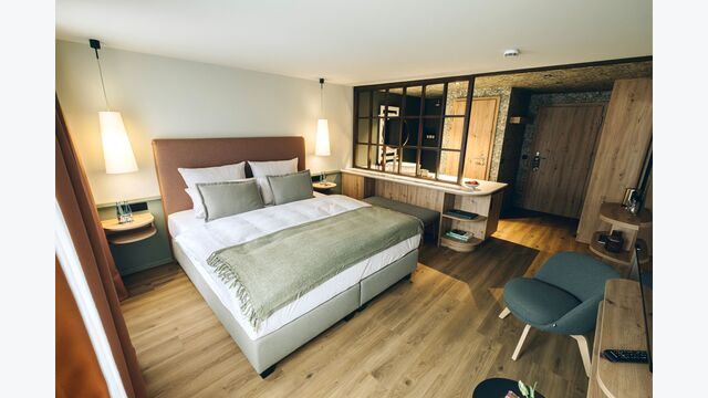 Das neu gestaltete Hotelzimmer – akzentuiert durch gezielte Kontraste wie das dunkle Fenstergitter zur hellen Holzverkleidung und die Grüntöne zur warmen Lichtstimmung.